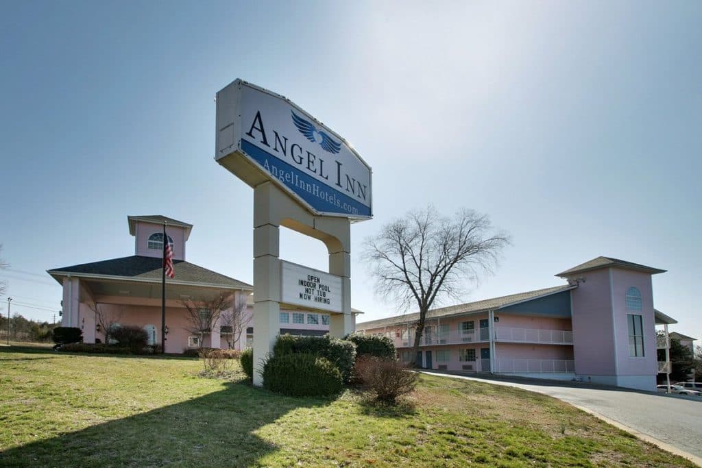 Exterior view of Angel Inn Central in Branson, MO with the hotel sign, pink buildings, covered entrance, and landscaped grounds on a sunny day
